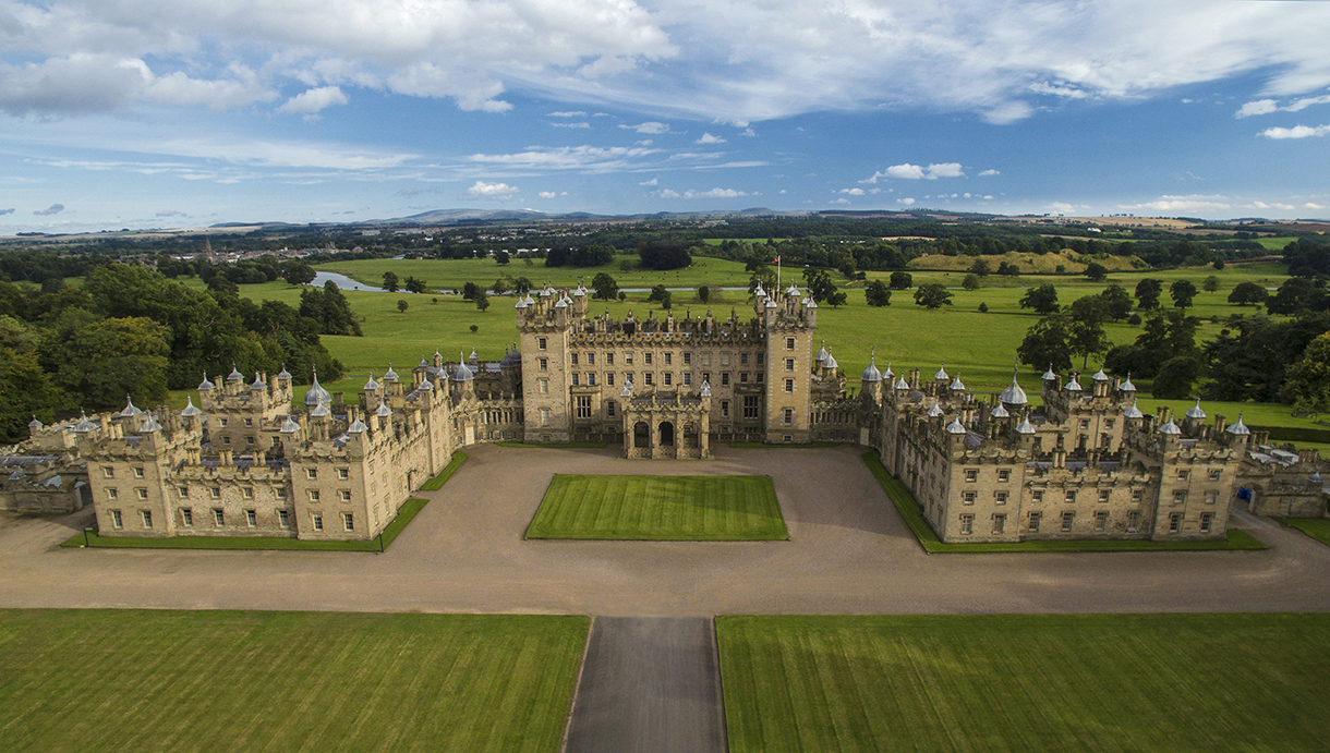 Exterior of Floors Castle located in the Scottish countryside. It is a grand castle with turrets, towers and a large central dome, situated in the midst of lush and manicured green gardens with trees and shrubs. 